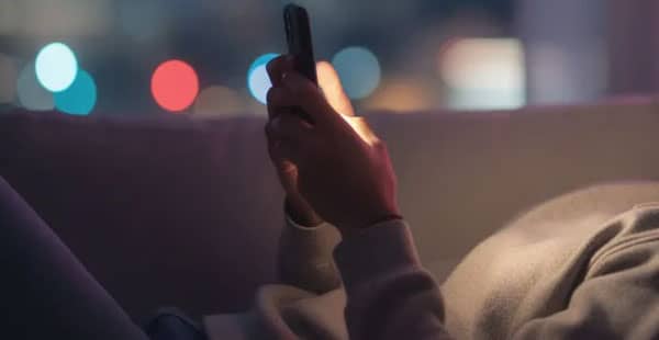 A person is seen relaxing while using a smartphone, surrounded by vibrant colorful lights in the background, creating a cozy atmosphere. This scene evokes a sense of enjoyment, reminiscent of the excitement found in online casinos and the thrill of playing casino games.